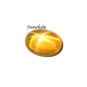 K&ouml;nnte beinhalten: Ein ovaler, goldgelber Edelstein mit einem ausgepr&auml;gten sechsstrahligen Sternmuster auf seiner glatten, polierten Oberfl&auml;che. Das Wort Snowwhite ist in schwarzer Schrift &uuml;ber dem Stein gedruckt, der einen durchscheinenden Cabochon-Schliff aufweist.