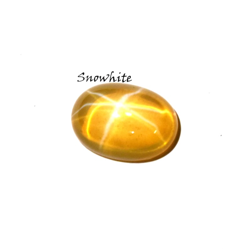 K&ouml;nnte beinhalten: Ein ovaler, goldgelber Edelstein mit einem ausgepr&auml;gten sechsstrahligen Sternmuster auf seiner glatten, polierten Oberfl&auml;che. Das Wort Snowwhite ist in schwarzer Schrift &uuml;ber dem Stein gedruckt, der einen durchscheinenden Cabochon-Schliff aufweist.