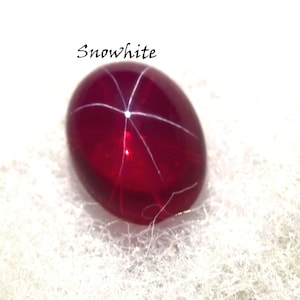 May include: A deep red, oval gemstone with a star-like pattern of white lines radiating from the centre. The word "Snowwhite" is written above the gem in a cursive font. The gem is set against a white, textured background.