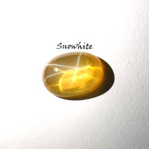 K&ouml;nnte beinhalten: Ein ovaler, durchscheinender gelber Edelstein mit einem ausgepr&auml;gten sechsstrahligen Sterneffekt, bekannt als Asterismus. Der Stein hat einen glatten, polierten Cabochon-Schliff und befindet sich unter dem Text Snowwhite in schwarzer Serifenschrift.