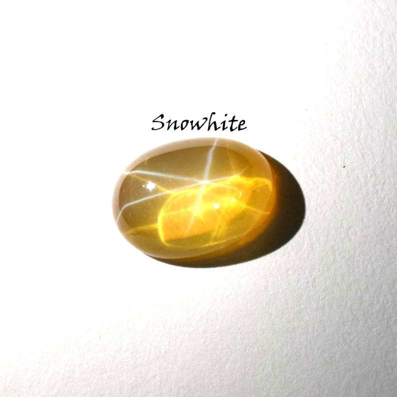 K&ouml;nnte beinhalten: Ein ovaler, durchscheinender gelber Edelstein mit einem ausgepr&auml;gten sechsstrahligen Sterneffekt, bekannt als Asterismus. Der Stein hat einen glatten, polierten Cabochon-Schliff und befindet sich unter dem Text Snowwhite in schwarzer Serifenschrift.