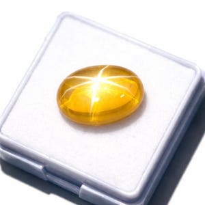 K&ouml;nnte beinhalten: Ein polierter, ovaler gelber Edelstein mit einem ausgepr&auml;gten sechsstrahligen Sterneffekt, bekannt als Asterismus. Der im Cabochon-Schliff gearbeitete Stein zeigt einen leuchtend goldenen Farbton und liegt in einer quadratischen wei&szlig;en Box.