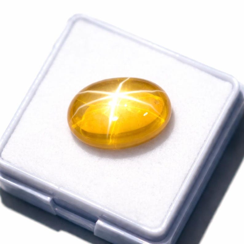 K&ouml;nnte beinhalten: Ein polierter, ovaler gelber Edelstein mit einem ausgepr&auml;gten sechsstrahligen Sterneffekt, bekannt als Asterismus. Der im Cabochon-Schliff gearbeitete Stein zeigt einen leuchtend goldenen Farbton und liegt in einer quadratischen wei&szlig;en Box.