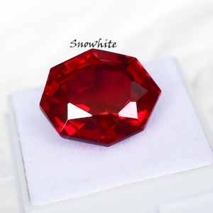 Könnte beinhalten: Ein facettierter, achteckiger, tiefroter Edelstein. Der Stein liegt auf einer weißen Oberfläche und trägt das Wort "Snowwhite" in Schreibschrift. Die Facetten des Steins reflektieren das Licht.