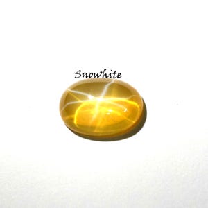 K&ouml;nnte beinhalten: Ein ovaler, durchscheinender gelber Edelstein mit einem ausgepr&auml;gten sechsstrahligen Sterneffekt, bekannt als Asterismus. Der Stein hat einen glatten, polierten Cabochon-Schliff und befindet sich unter dem Text Snowwhite in schwarzer Schreibschrift.