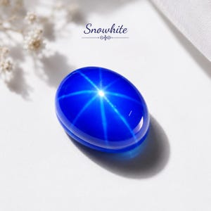 Puede incluir: Una piedra preciosa azul pulida de forma ovalada que presenta un distintivo efecto de estrella de seis rayos en su superficie. La piedra es de color azul intenso con un acabado liso y reflectante. El texto Snowwhite aparece en azul oscuro.