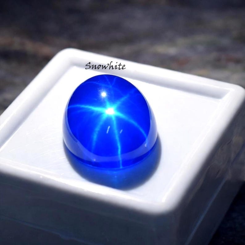 Puede incluir: Una gema cabuj&oacute;n ovalada de color azul vibrante con un efecto de estrella de seis puntas, centrada sobre una base de exhibici&oacute;n cuadrada blanca. La palabra Snowhite est&aacute; impresa en texto negro sobre la gema en la caja de exhibici&oacute;n.