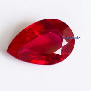 May include: A faceted, pear-shaped ruby gemstone. The gem is a deep, vibrant red colour and has a polished, reflective surface. The word "Snowwhite" is written in blue script on the right side of the gem.