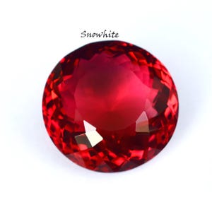 May include: A faceted, round, red gemstone with the word "Snowwhite" in cursive script above it. The gem has a brilliant cut, reflecting light and creating a sparkling effect. The gem is set against a white background.
