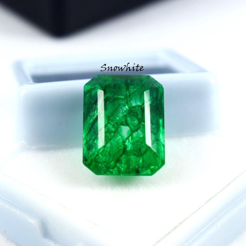 May include: A vibrant, emerald-cut green gemstone with the word "Snowwhite" printed above it. The gem is set against a white surface and a light blue background. The gemstone's facets reflect light, showcasing its clarity and color.