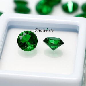May include: Two faceted, round, emerald green gemstones are displayed in a white square container, with the word "Snowwhite" above them. Several other green gemstones are scattered around the container, creating a sparkling effect.