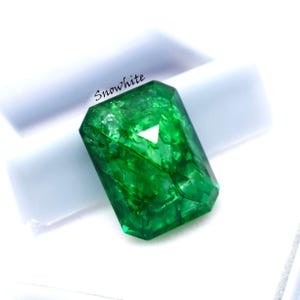 May include: A faceted, emerald-green gemstone with a rectangular shape. The gem is set against a white background and has the word "Snowwhite" printed on it. The gem's facets reflect light, creating a sparkling effect.