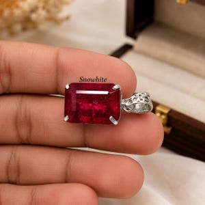 Könnte beinhalten: Ein rechteckiger, tiefroter Edelsteinanhänger in einer silberfarbenen Metallfassung mit einer dekorativen filigranen Öse. Das Wort Snowwhite ist in schwarzer Schrift über dem facettierten Edelstein aufgedruckt.