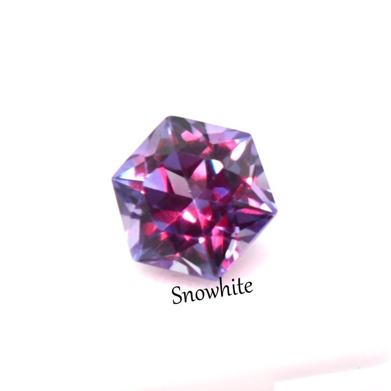 May include: A faceted hexagonal gemstone displaying deep purple and magenta hues with reflective surfaces. The stone features a brilliant cut that creates sharp geometric light patterns. The word Snowwhite is printed in black text below the gem.