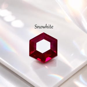 Può includere: Una gemma esagonale sfaccettata di colore rosso intenso, con un centro esagonale bianco. La parola "Snowwhite" è stampata sopra la gemma. La gemma è posta su una superficie bianca.