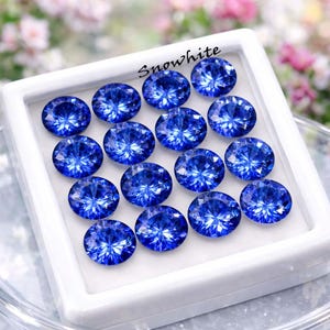 Op de afbeelding: Een verzameling ronde, gefacetteerde, briljantblauwe edelstenen, gerangschikt in een witte vitrine. De edelstenen zijn zeer reflecterend en vangen het licht op, waardoor een sprankelend effect ontstaat. Het woord "Snowwhite" staat cursief geschreven.