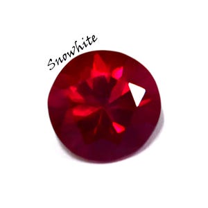Peut inclure: Une pierre précieuse rouge foncé, ronde et facettée, avec le mot "Snowwhite" en écriture cursive. La gemme a une taille brillante, reflétant la lumière. Le fond est blanc.