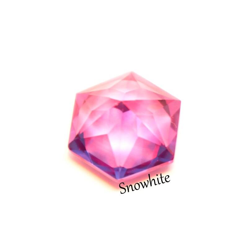May include: A faceted hexagonal gemstone with a vibrant pink and purple gradient color scheme. The stone features a geometric cut with multiple triangular surfaces that reflect light. The word Snowwhite is printed in black text below the stone.