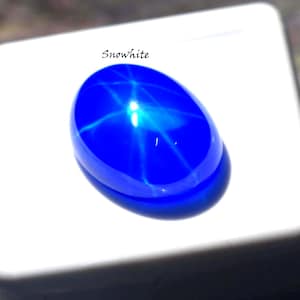 Puede incluir: Una piedra preciosa cabuj&oacute;n de color azul vibrante y forma ovalada que presenta un efecto de estrella de seis rayos en su superficie pulida. La palabra Snowhite est&aacute; impresa en negro sobre la piedra, que descansa en una base blanca.