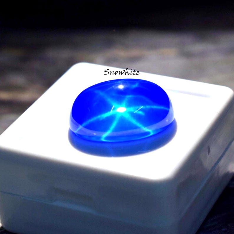 Puede incluir: Una gema cabuj&oacute;n de color azul vibrante que presenta un efecto de estrella de seis puntas en su superficie. La piedra se muestra sobre una base cuadrada blanca con la palabra Snowhite impresa en texto negro sobre la gema, mostrando su acabado.