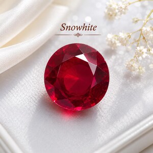 May include: A large, round, faceted ruby gemstone with a deep red color. The gemstone is set against a white satin background, with the word "Snowwhite" above it. Delicate white flowers are in the background.