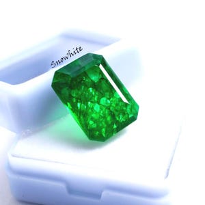 May include: A vibrant, emerald-cut green gemstone rests in a white box. The gem has a faceted surface, reflecting light and showcasing its clarity. The word "Snowwhite" is printed on the box. This image is ideal for jewelry and gemstone listings.