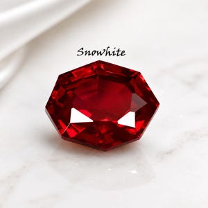 Op de afbeelding: Een gefacetteerde, zeshoekige dieprode edelsteen met een briljant slijpvorm. Het woord "Snowhite" staat in zwarte schrijfletters boven de steen gedrukt. De edelsteen vertoont scherpe, geometrische randen en een levendige, verzadigde karmozijnrode kleur.