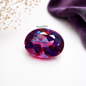 May include: A large, oval-cut gemstone displaying a spectrum of colours, predominantly purple, pink, and red. The gemstone is set against a white backdrop, with the word "Snowwhite" above it. A purple fabric is in the background.