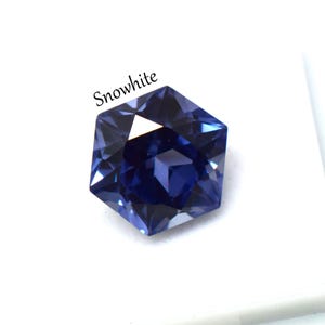 May include: A faceted hexagonal gemstone in a deep royal blue color with a polished finish. The word Snowwhite is printed in black text above the stone. The gem features multiple triangular facets that reflect light, creating a sharp geometric shape.