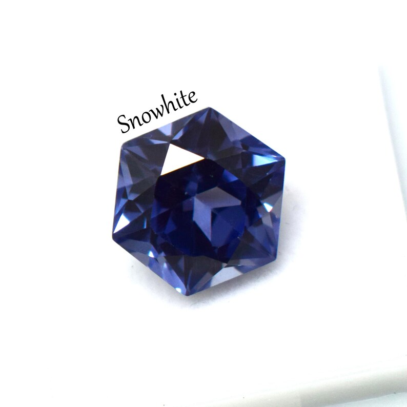 May include: A faceted hexagonal gemstone in a deep royal blue color with a polished finish. The word Snowwhite is printed in black text above the stone. The gem features multiple triangular facets that reflect light, creating a sharp geometric shape.