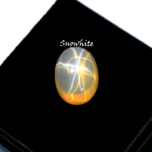 K&ouml;nnte beinhalten: Ein ovaler, durchscheinender wei&szlig;er Cabochon-Edelstein mit einem ausgepr&auml;gten sechsstrahligen Sterneffekt auf der Oberfl&auml;che. Der Stein ist mit dem Text Snowwhite beschriftet und weist eine glatte, polierte Oberfl&auml;che sowie ein sanftes Leuchten auf.