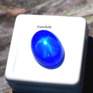 Puede incluir: Una gema ovalada de color azul vibrante en cabuj&oacute;n que presenta un efecto de estrella de seis puntas centrado en su superficie. La gema se muestra sobre una base cuadrada blanca con la palabra Snowhite impresa en negro arriba.