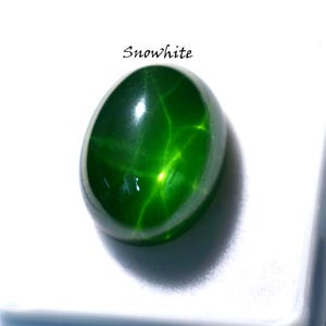 Pode incluir: Uma gema verde, de forma oval, com um padrão em forma de estrela. A gema é altamente polida e reflete a luz, criando um efeito luminoso. A palavra "Snowwhite" está escrita acima da gema.