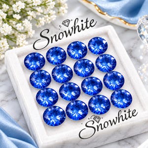 May include: A collection of fifteen round, faceted blue gemstones arranged in a grid within a white square display tray. The text Snowwhite is printed in black script above and below the stones, accompanied by a small diamond icon.