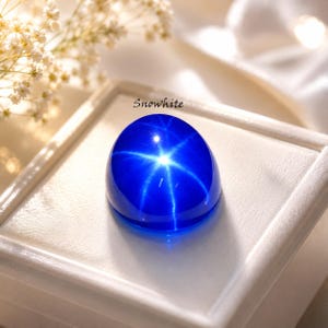 Puede incluir: Una gema cabuj&oacute;n de color azul vibrante que presenta un efecto de estrella de seis rayos, conocido como asterismo, centrado en su superficie lisa y pulida. La piedra tiene la inscripci&oacute;n Snowwhite y descansa sobre una base de exhibici&oacute;n blanca.