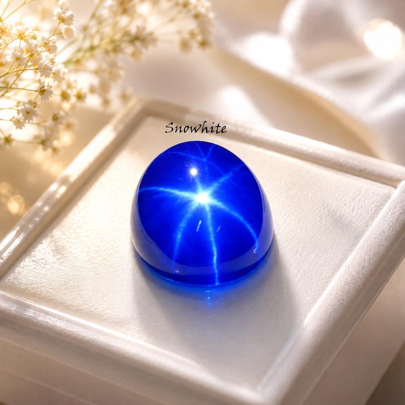 Puede incluir: Una gema cabuj&oacute;n de color azul vibrante que presenta un efecto de estrella de seis rayos, conocido como asterismo, centrado en su superficie lisa y pulida. La piedra tiene la inscripci&oacute;n Snowwhite y descansa sobre una base de exhibici&oacute;n blanca.