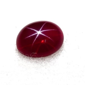 May include: A polished, oval-shaped, deep red gemstone with a six-rayed star pattern. The star effect is created by light reflecting off inclusions within the stone. The gem is set against a white background.