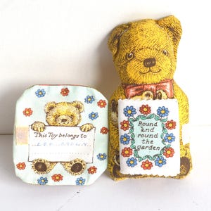May include: A plush yellow teddy bear with a red bow tie holds a sign that reads "Round and round the garden" surrounded by flowers. Beside it is a matching fabric box with a teddy bear graphic and the text "This Toy belongs to".
