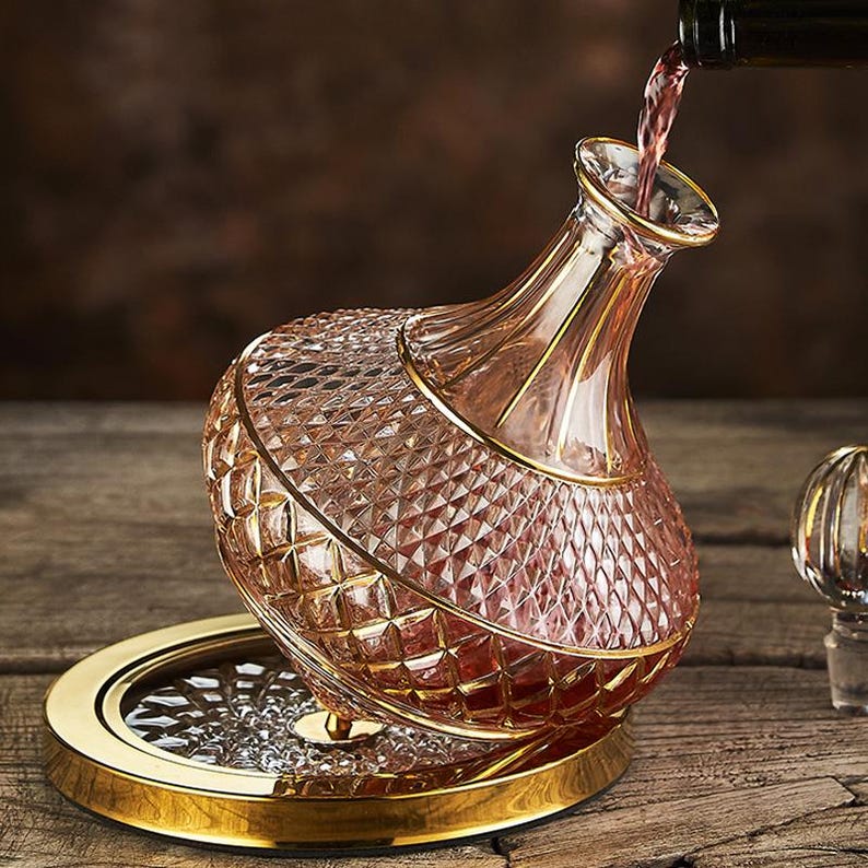 May include: A crystal wine decanter with a diamond-cut pattern and gold trim is being filled with red wine. The decanter sits on a gold-rimmed base. The wine is being poured from a dark bottle.
