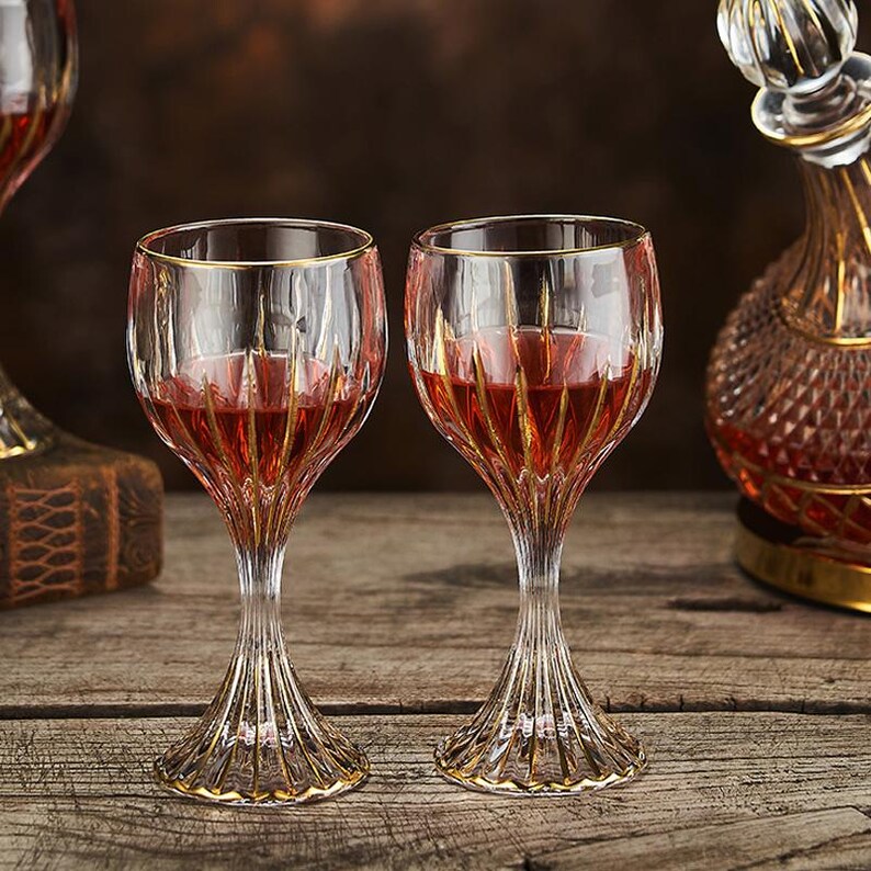 May include: Two clear wine glasses with gold accents, filled with red liquid, sit on a wooden surface. The glasses have a detailed, ribbed design and a gold rim. A decanter with a gold base and stopper is visible in the background.