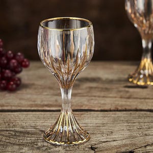 May include: Clear wine glass with gold trim. The glass has a textured pattern and a gold rim. The stem and base are also accented with gold. The glass is on a wooden surface.