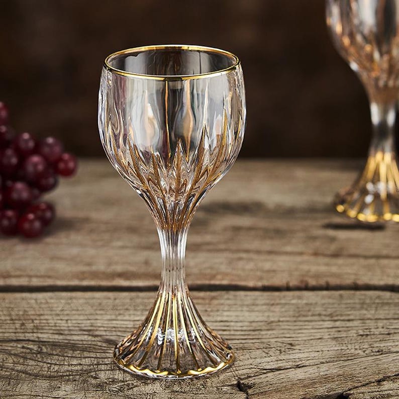 May include: Clear wine glass with gold trim. The glass has a textured pattern and a gold rim. The stem and base are also accented with gold. The glass is on a wooden surface.