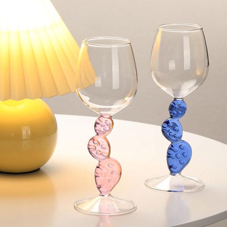 May include: Two clear wine glasses with unique stems. One stem is pink, the other is blue, both with a stacked, textured design. The glasses sit on a clear base, and the bowl of the glass is clear.