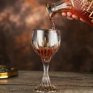 May include: A clear wine glass with gold accents is being filled with red wine from a matching decanter. The glass has a long, elegant stem and a detailed design. The decanter has a gold rim and a diamond pattern.