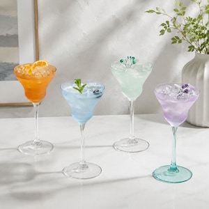 May include: Four stemmed cocktail glasses filled with ice and colorful beverages. The glasses contain orange, light blue, pale green, and light purple liquids, garnished with orange slices, mint leaves, blueberries, and small floral sprigs.