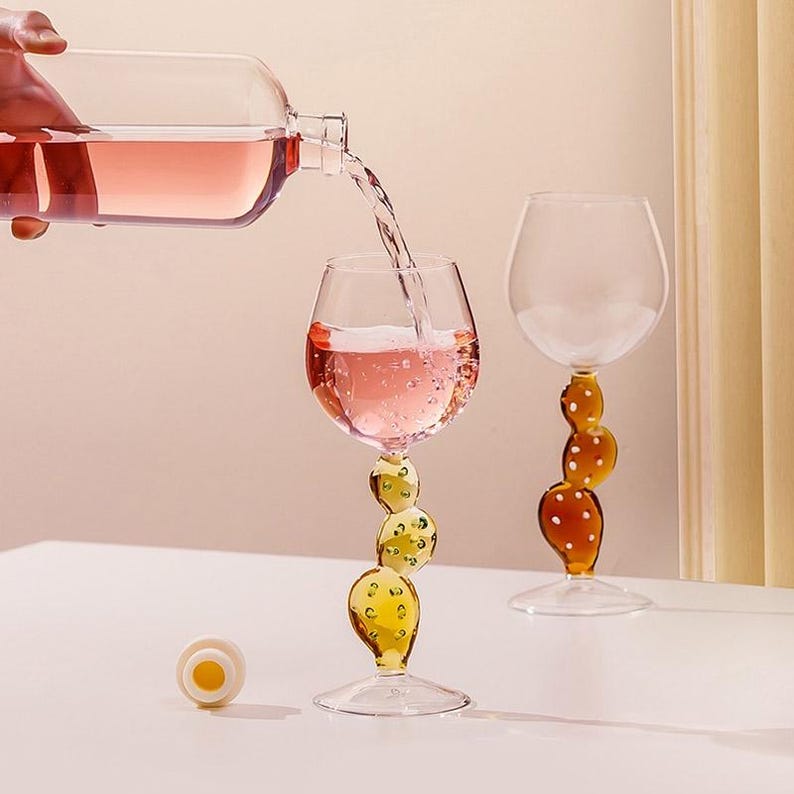 May include: A clear wine glass is filled with pink liquid as it is poured from a bottle. The glass has a yellow stem with a bubble design. Another glass with a brown stem and a bubble design is next to it. A white and yellow bottle stopper is on the table.