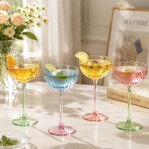 Può includere: Quattro coppe da cocktail con coppe scanalate e basi in vetro colorato. I bicchieri contengono cocktail colorati guarniti con fette di agrumi e menta. Il primo bicchiere è giallo con base verde, il secondo blu con base rosa, il terzo giallo con base rosa e il quarto rosa con base verde.