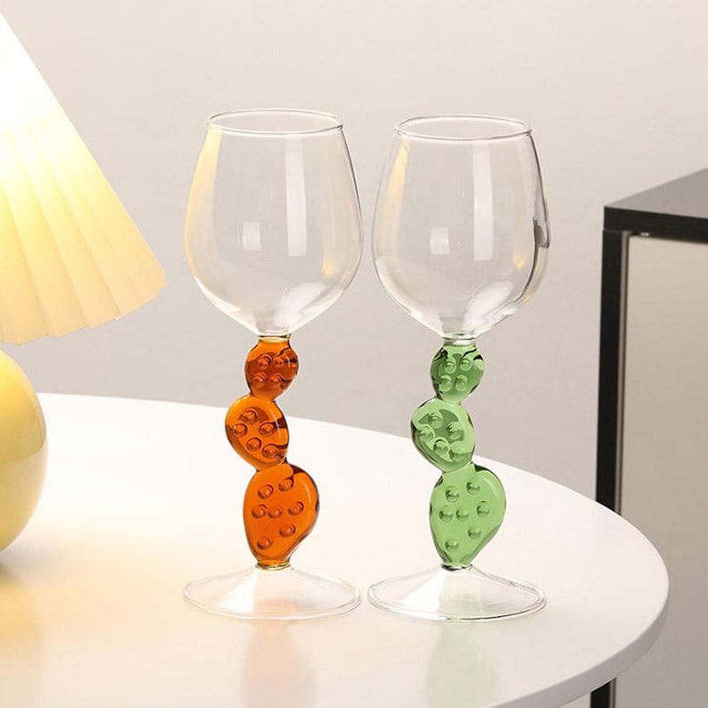 May include: Two clear wine glasses with unique stems. One stem is orange, the other is green. Each stem features a stacked, textured design. The glasses sit on a white surface, with a lamp and cabinet in the background.