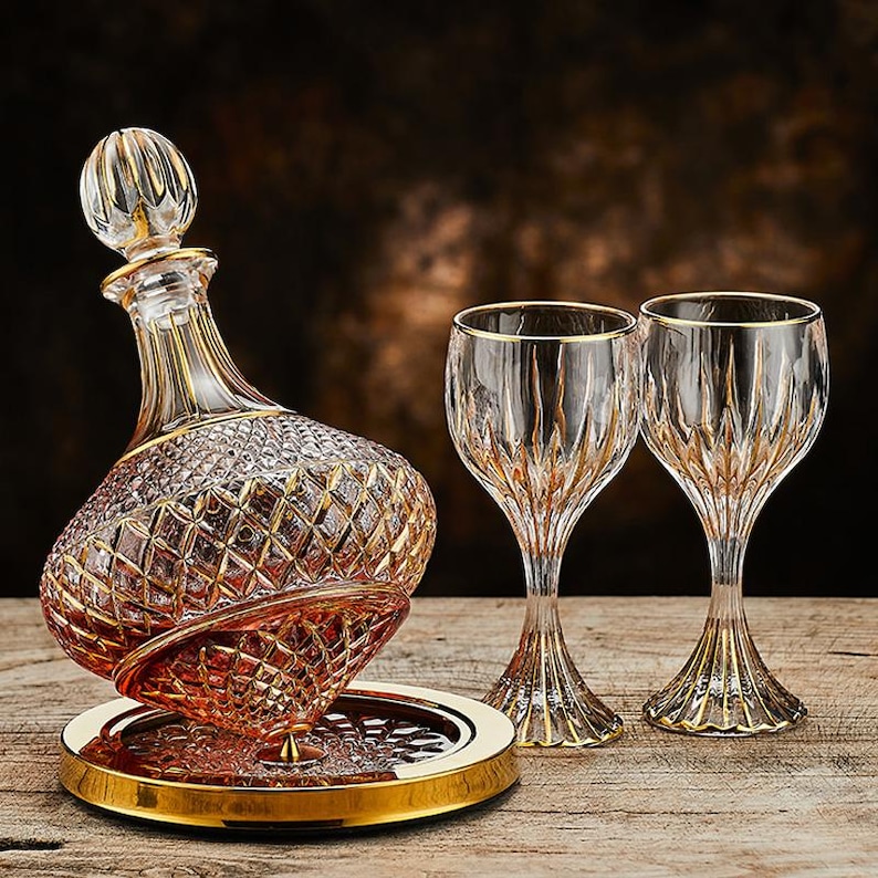 May include: A crystal decanter set with two matching wine glasses. The decanter has a diamond pattern with gold trim, and a gold-trimmed stopper. The glasses and decanter sit on a gold-rimmed tray, all on a wooden surface.