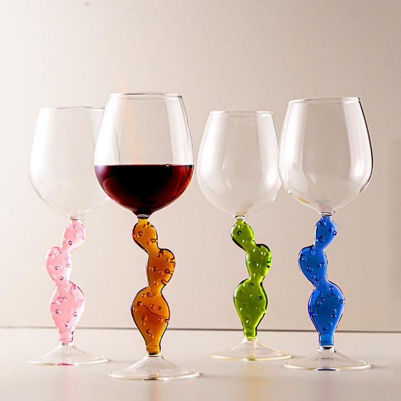 May include: Four clear wine glasses with colorful cactus-shaped stems. One glass is filled with red wine. The stems are pink, orange, green, and blue. The glasses are on a white surface.